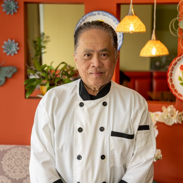Smiling Asian chef in a white uniform with black trim, standing in a restaurant.
