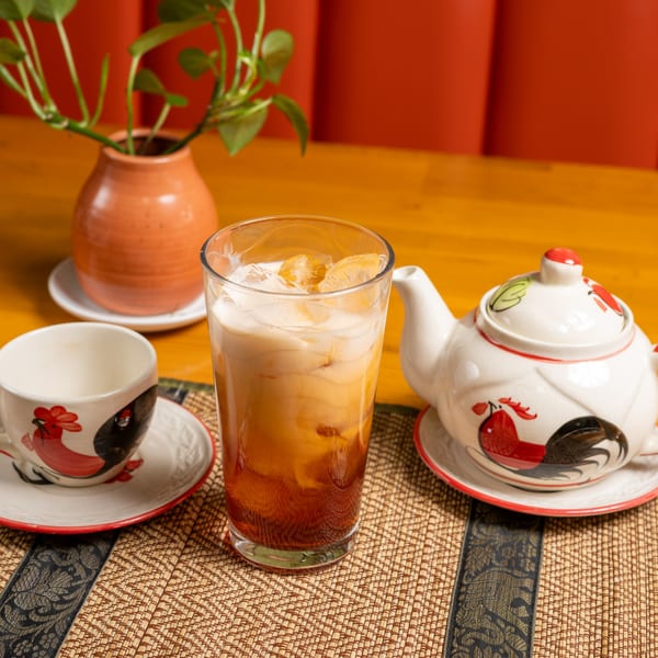 Iced Thai tea with a teapot and cup featuring rooster designs on a woven mat.