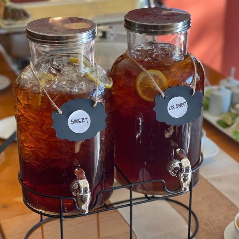 Two glass dispensers of iced tea, one sweet and one unsweet, with lemon slices and ice.