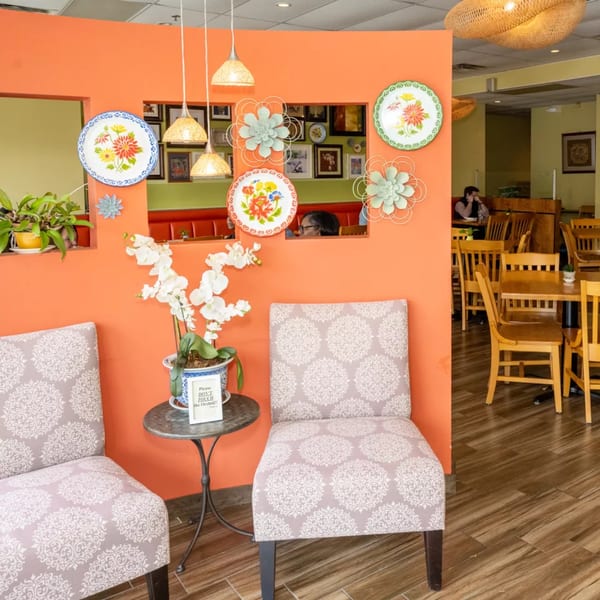 Restaurant interior with orange walls, decorative plates, potted orchids, and patrons dining at wooden tables.