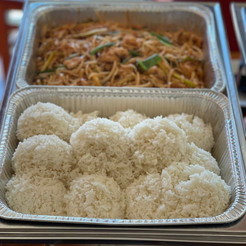 Two foil trays of food, one with white rice and the other with noodles and vegetables.