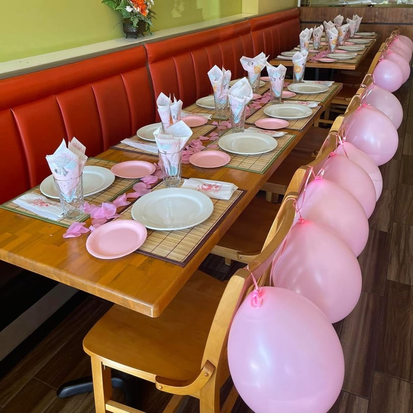 Restaurant tables set with pink plates, napkins, and pink balloons tied to chairs for a party.