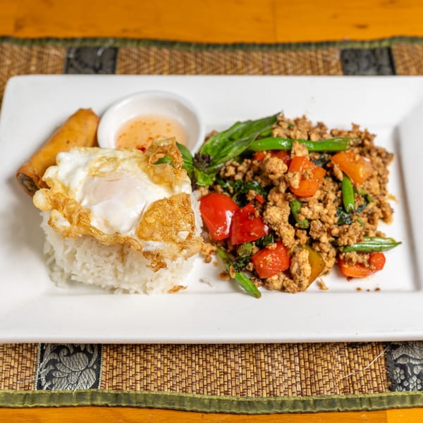 Thai basil chicken with rice, fried egg, spring roll, and dipping sauce on a white plate.