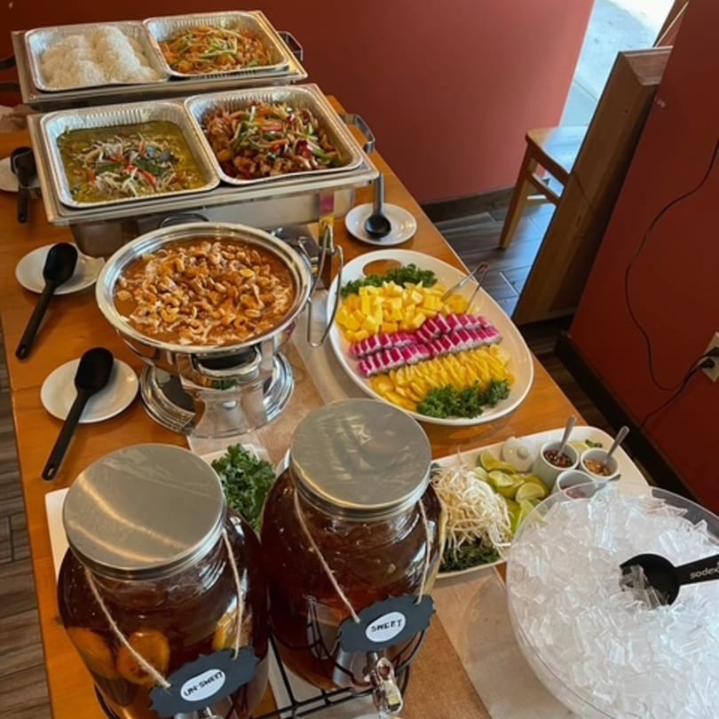 A buffet table laden with Thai dishes, including curries, noodles, rice, fresh fruit, and iced tea.