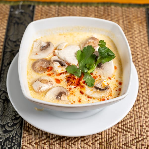 Creamy Tom Kha Gai soup with mushrooms, chicken, and cilantro in a white bowl on a woven mat.