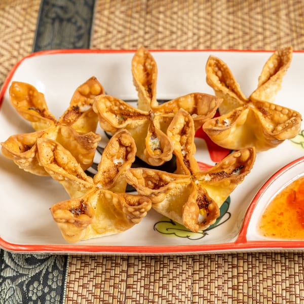 Golden-brown crab rangoon appetizers, shaped like flowers, served with sweet chili dipping sauce on a white plate.